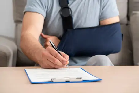 Person with an injured arm in a sling signing paperwork while seated at a table. | John Michael Morrow Law Firm